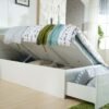 A white Adela King Single Storage Bed shown open, with its gas-lift mattress base raised to reveal a large under-bed storage compartment, alongside a fully extended footend drawer with striped pillows inside, in a modern bedroom setting.