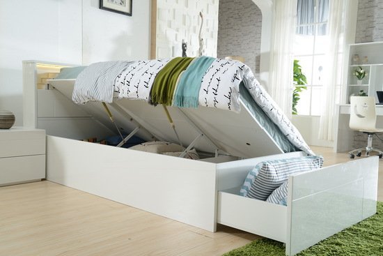A white Adela King Single Storage Bed shown open, with its gas-lift mattress base raised to reveal a large under-bed storage compartment, alongside a fully extended footend drawer with striped pillows inside, in a modern bedroom setting.