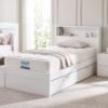 A high-quality photo of a modern white Adela King Single bed in a naturally lit bedroom. Features include a quilted SleepMaker mattress, a built-in shelving headboard with books, a matching 2-drawer nightstand, and visible pull-out fabric handles for a trundle/storage drawer on its white base.